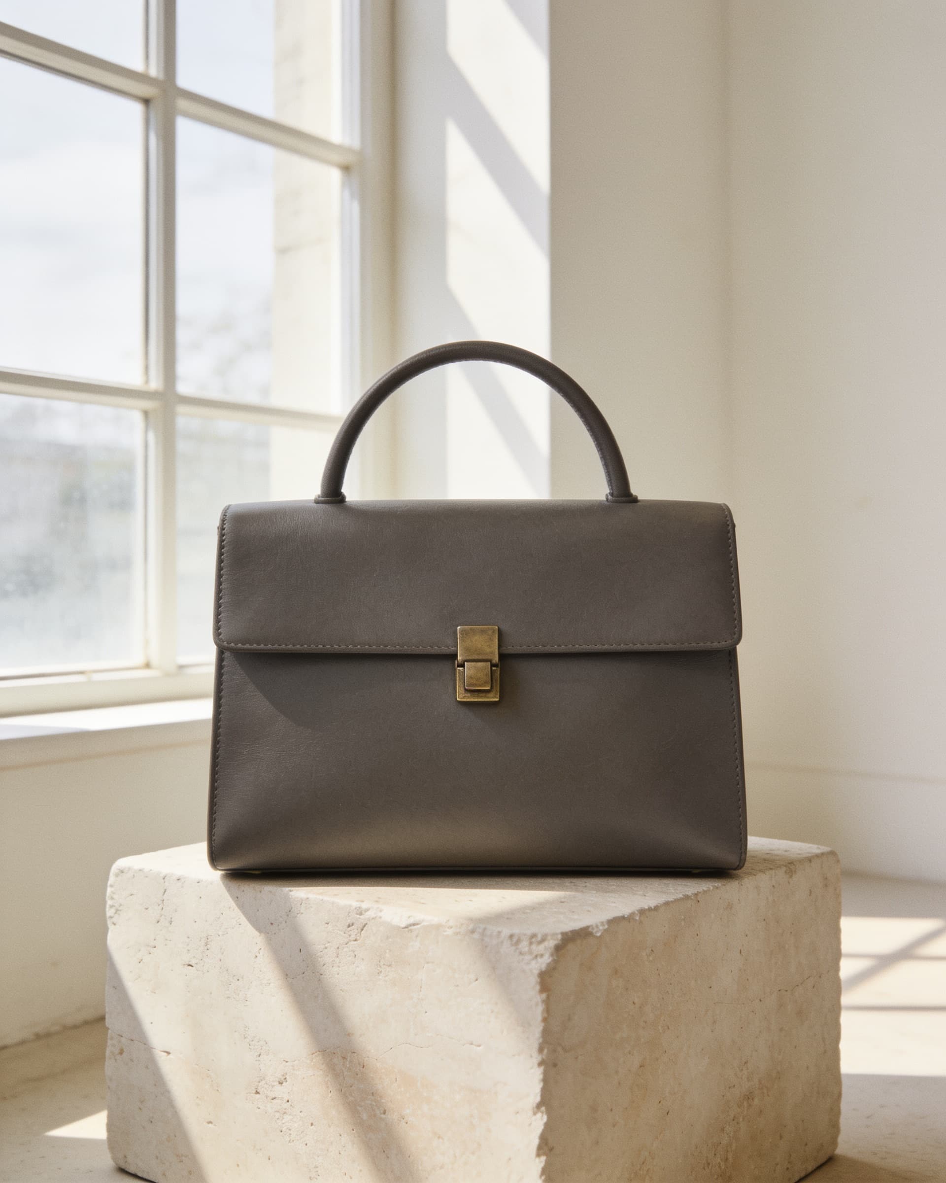 Bolso de piel charcoal de líneas rectas con cierre de latón, sobre un bloque de piedra